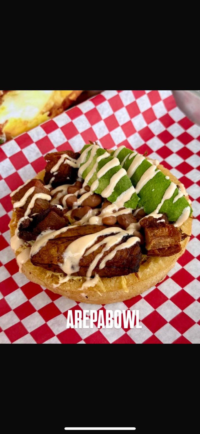 Arepabowl with plantains, beans, avocado slices, and creamy sauce on red and white checkered paper.