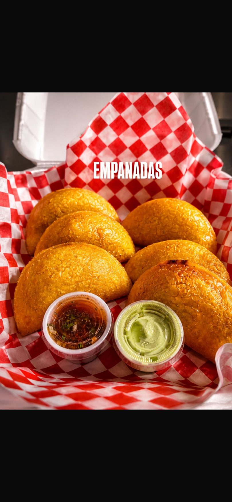 Golden-brown empanadas and two dipping sauces in a red and white checkered paper-lined container.