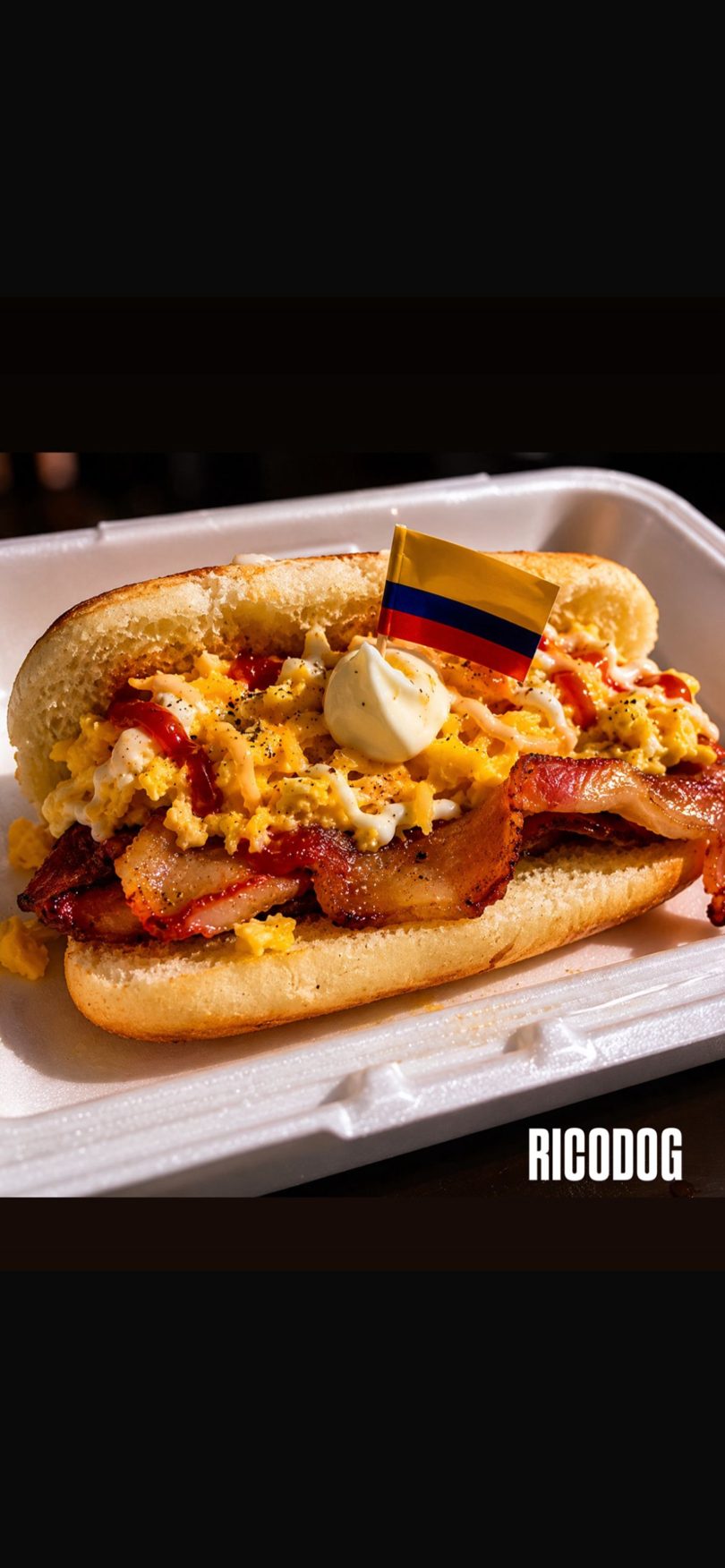A Ricodog hot dog with scrambled eggs, bacon, and sauces, topped with a Colombian flag.
