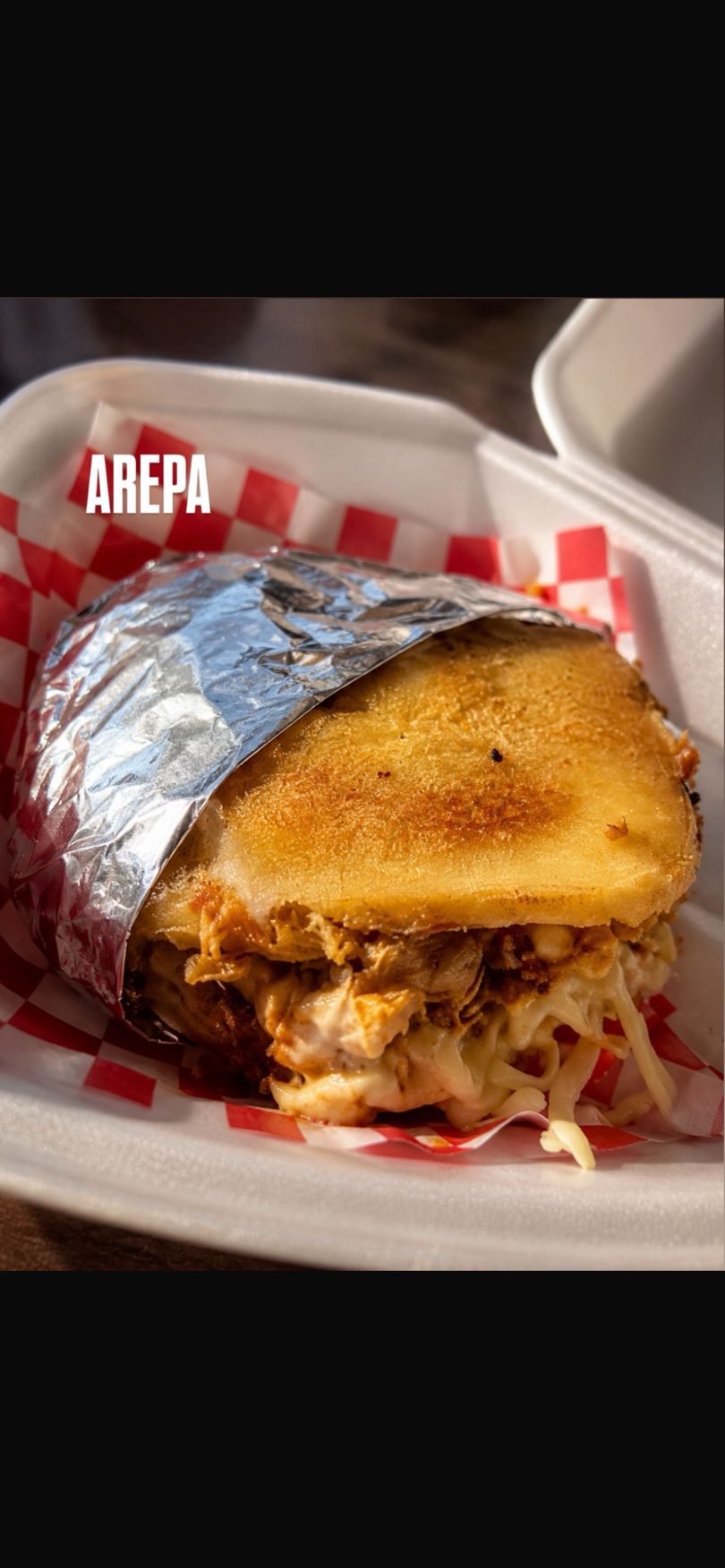 A golden-brown arepa, partially wrapped in foil, filled with shredded chicken and cheese.
