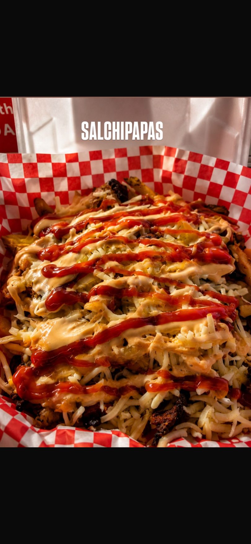 Salchipapas, a dish of fries topped with cheese, ketchup, and a creamy sauce in a red and white checkered basket.